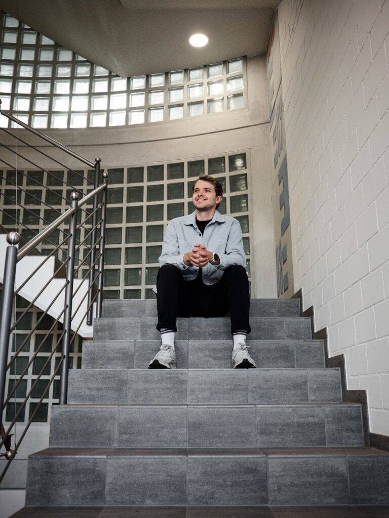 Junger Mann lächelnd sitzt auf Treppe vor Glasbausteinwand, modernes Interieur, Fotograf Winterthur