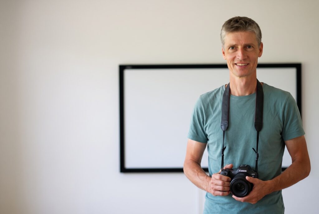 Lächelnder Fotograf mit Canon vor heller Wand; Fotostudio, Fotograf Winterthur, Videoproduktion